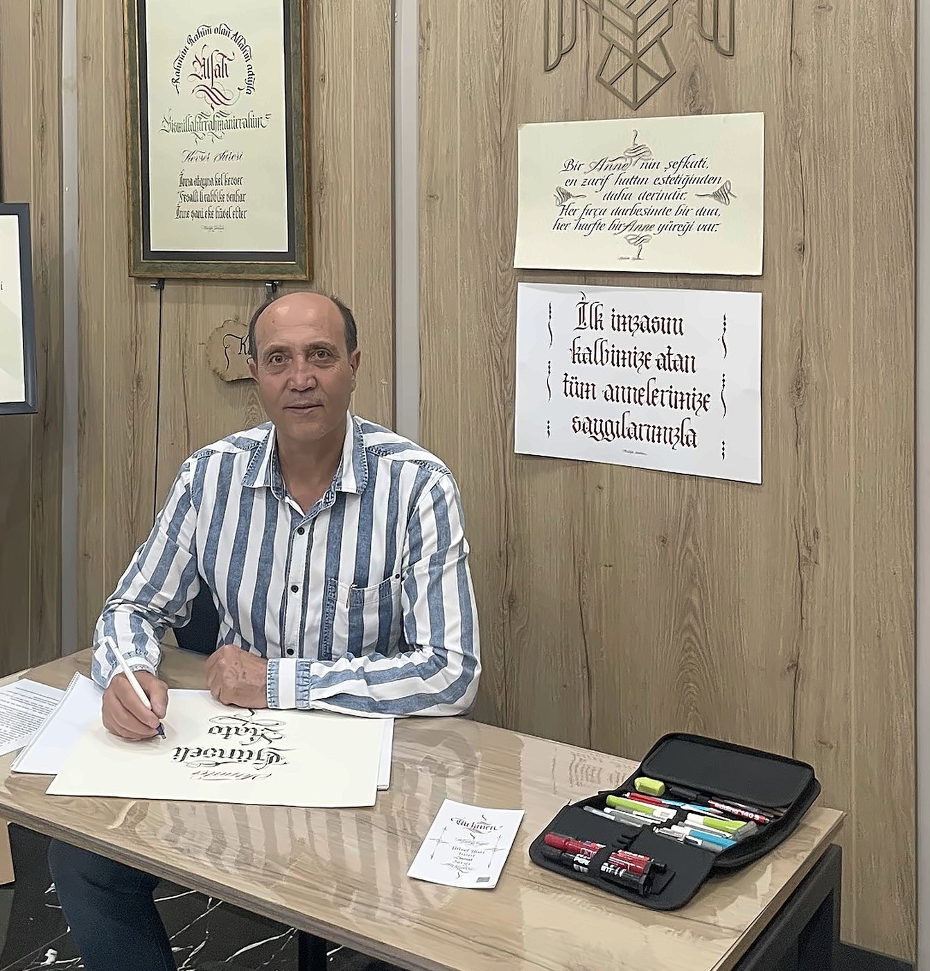Mustafa Türkmen atölyesinde, arkasında çerçeveli kaligrafi eserleriyle masasında yazarken