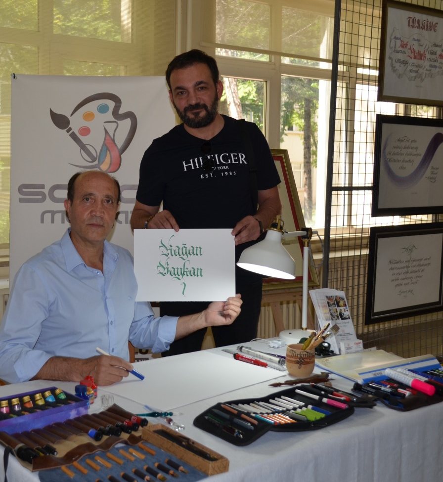 Mustafa Türkmen, masasında; yanındaki ziyaretçi onun yazdığı ismi gösteriyor
